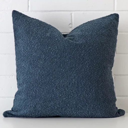 Square cushion cover in blue colour sitting upright in front of a brick wall. It has been made from a quality boucle material.