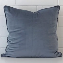 An attractive velvet cushion in front of a white brick wall. It has a square shape and is blue grey in colour.