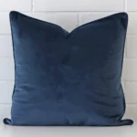 Blue cushion cover sits against a white wall. It is constructed from a superior looking velvet material and has square dimensions.