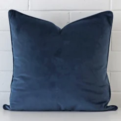 Blue cushion cover sits against a white wall. It is constructed from a superior looking velvet material and has square dimensions.