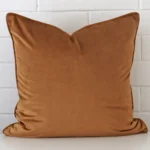 Velvet square cushion in an upright position against a white brick wall. It is bronze in colour.