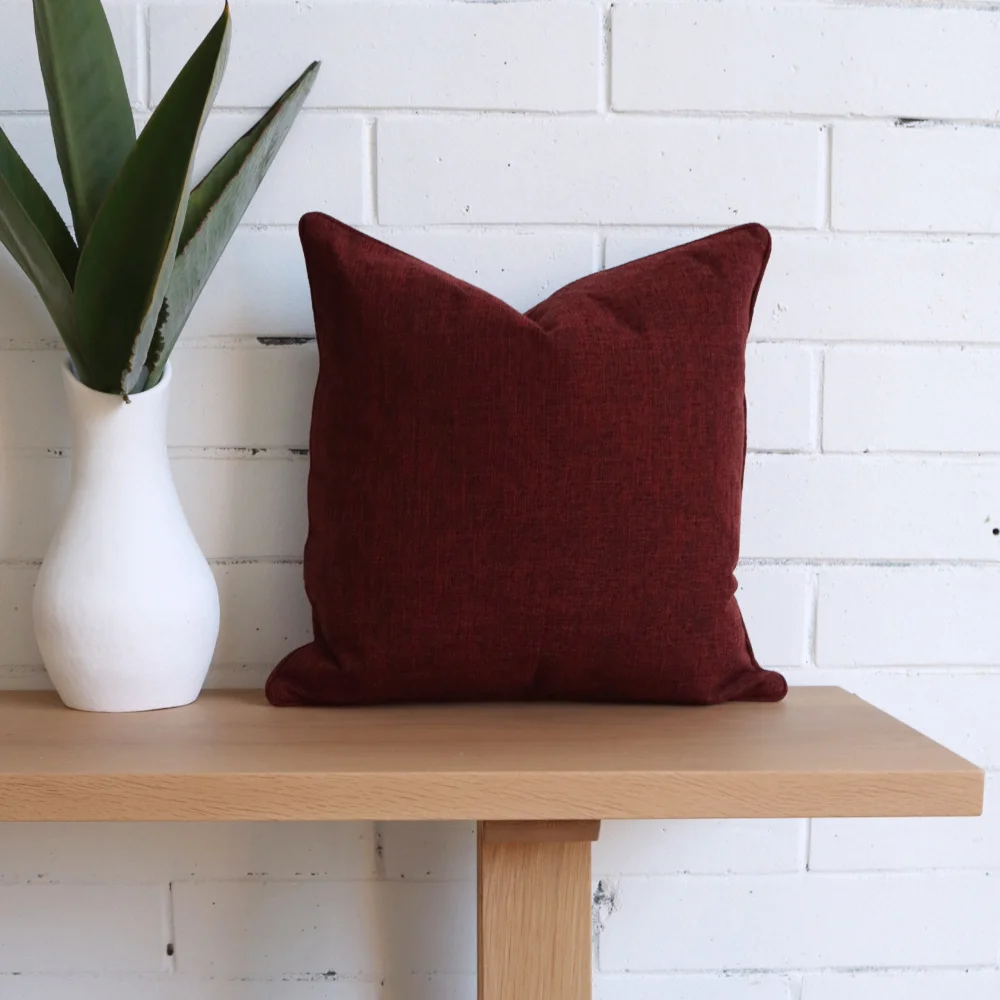 A burgundy cushion is laying against a white brick wall on a seat and next to a vase.