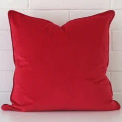 velvet square cushion in an upright position against a white brick wall. It is red in colour.