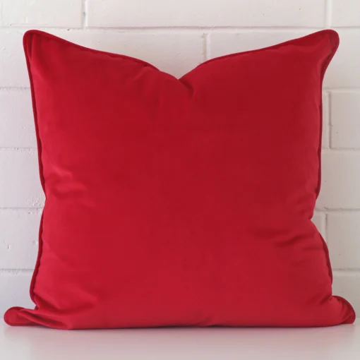 velvet square cushion in an upright position against a white brick wall. It is red in colour.