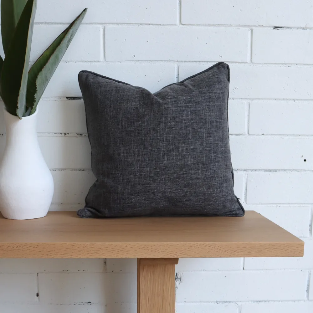 A charcoal cushion is sitting upright on a wooden bench and against a white wall