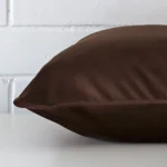 Chocolate brown cushion cover laying sideways against brick wall. The square size and velvet material are shown highlighting the seams.