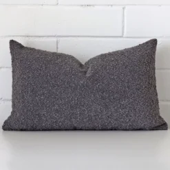 Dark grey cushion cover sits against a white wall. It is constructed from a superior looking boucle material and has rectangle dimensions.