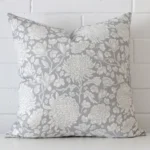 An attractive floral linen cushion in front of a white brick wall. It has a square shape and is ligt blue in colour.