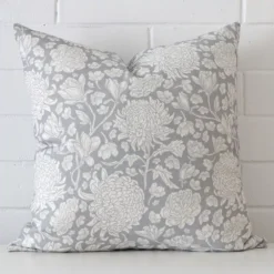 An attractive floral linen cushion in front of a white brick wall. It has a square shape and is ligt blue in colour.