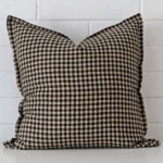 Designer square cushion with a gingham design in an upright position against a white brick wall.