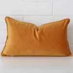 An attractive velvet cushion in front of a white brick wall. It has a rectangle shape and is gold mustard in colour.