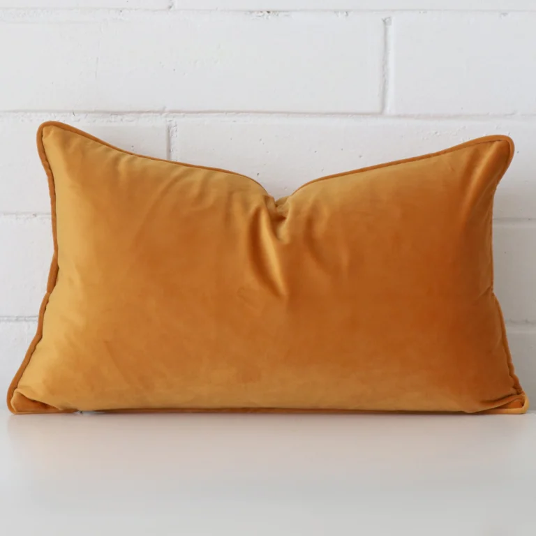 An attractive velvet cushion in front of a white brick wall. It has a rectangle shape and is gold mustard in colour.