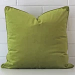 Green velvet cushion cover in front of a white wall. It has a square size and is made from a velvet material.
