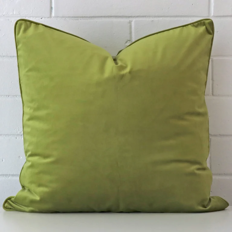 Green velvet cushion cover in front of a white wall. It has a square size and is made from a velvet material.