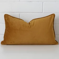 White wall with a honey mustard cushion laying against it. It has a distinctive velvet fabric and has a rectangle shape.