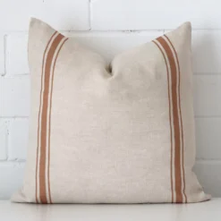 An attractive striped linen cushion in front of a white brick wall. It has a square shape and is terracotta in colour.