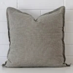 Bold square cushion positioned in front of white brickwork. Its gingham style pops on the designer fabric.