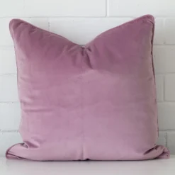 An attractive velvet cushion in front of a white brick wall. It has a square shape and is lavender in colour.