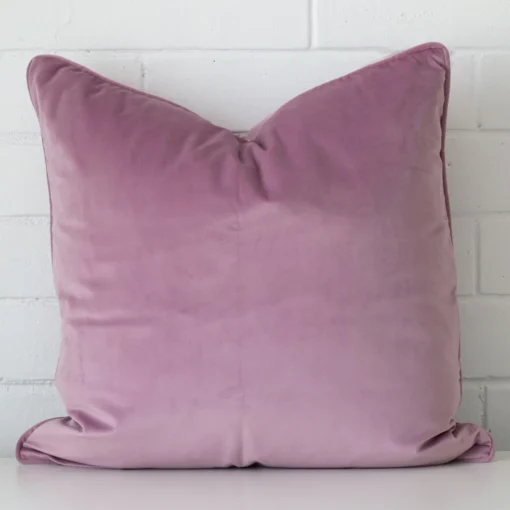 An attractive velvet cushion in front of a white brick wall. It has a square shape and is lavender in colour.