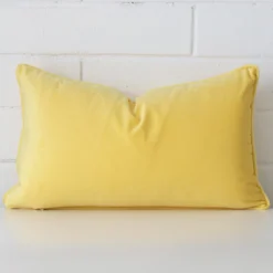 Velvet rectangle cushion in an upright position against a white brick wall. It is lemon yellow in colour.