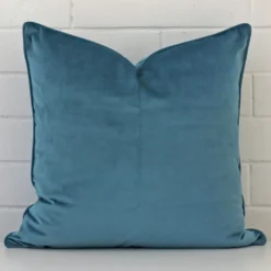 White wall with a light teal cushion laying against it. It has a distinctive velvet fabric and has a square shape.