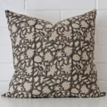 Square cushion with a floral design in an upright position against a white brick wall. It has been made from a quality designer material.