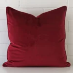 Velvet square cushion in an upright position against a white brick wall. It is maroon in colour.