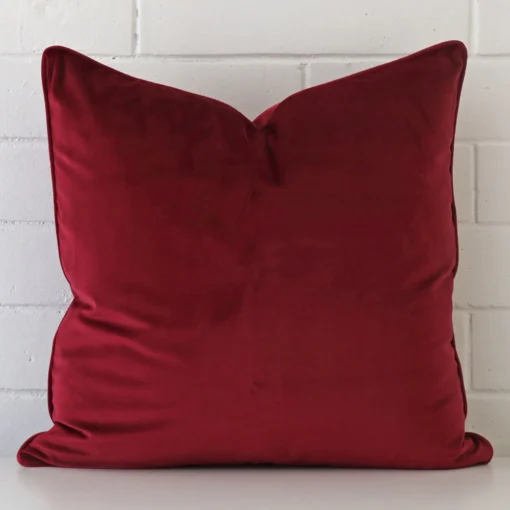 Velvet square cushion in an upright position against a white brick wall. It is maroon in colour.