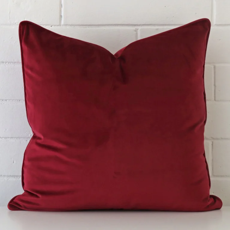 Velvet square cushion in an upright position against a white brick wall. It is maroon in colour.