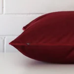 Maroon cushion cover laying sideways against brick wall. The square size and velvet material are shown highlighting the seams.