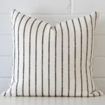 Striped cushion cover sits against a white wall. It is constructed from a superior looking linen material and has square dimensions.