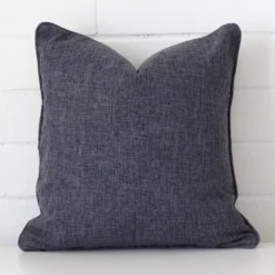 White wall with a charcoal cushion laying against it. It has a distinctive linen fabric and has a square shape.