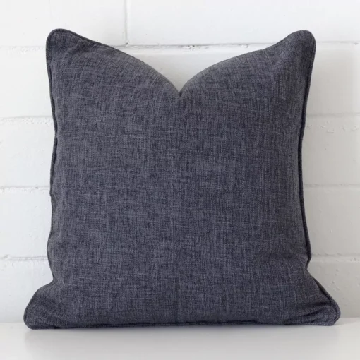 White wall with a charcoal cushion laying against it. It has a distinctive linen fabric and has a square shape.