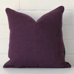 Square cushion cover in plum colour sitting upright in front of a brick wall. It has been made from a quality linen material.