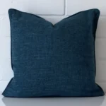 Linen square cushion in an upright position against a white brick wall. It is prussian blue in colour.