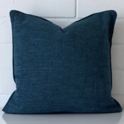 Linen square cushion in an upright position against a white brick wall. It is prussian blue in colour.