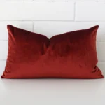 An attractive velvet cushion in front of a white brick wall. It has a rectangle shape and is rust in colour.