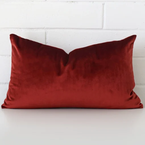 An attractive velvet cushion in front of a white brick wall. It has a rectangle shape and is rust in colour.