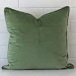 Bold large sage cushion positioned in front of white brickwork. It is made from velvet fabric.