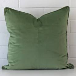 Bold large sage cushion positioned in front of white brickwork. It is made from velvet fabric.