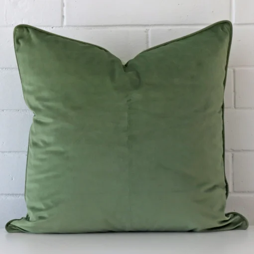 Bold large sage cushion positioned in front of white brickwork. It is made from velvet fabric.