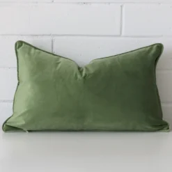 White wall with a sage cushion laying against it. It has a distinctive velvet fabric and has a rectangle shape.