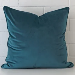 An attractive velvet cushion in front of a white brick wall. It has a large size and is teal in colour.