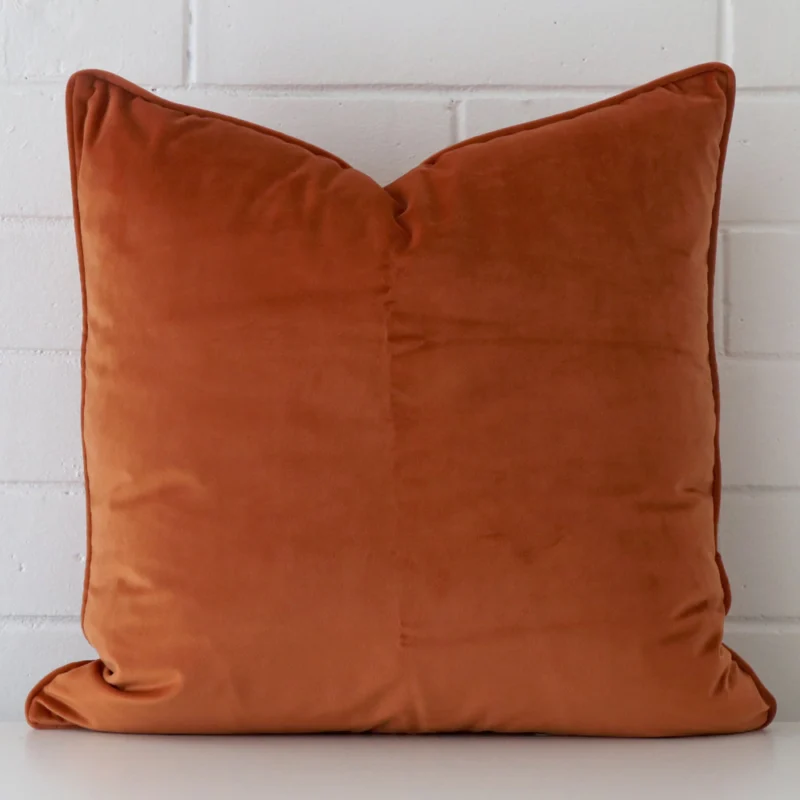 terracotta-velvet-cushion-cover-55cm-x-55cm-sc770-55b Velvet large cushion in an upright position against a white brick wall. It is terracotta in colour.