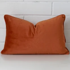 Velvet rectangle cushion in an upright position against a white brick wall. It is terracotta in colour.