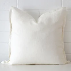 Bold square white cushion positioned in front of white brickwork. It has linen fabric.