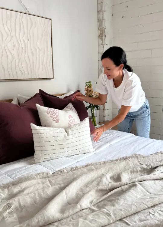 A woman is arranging four cushions in a bedroom setting.