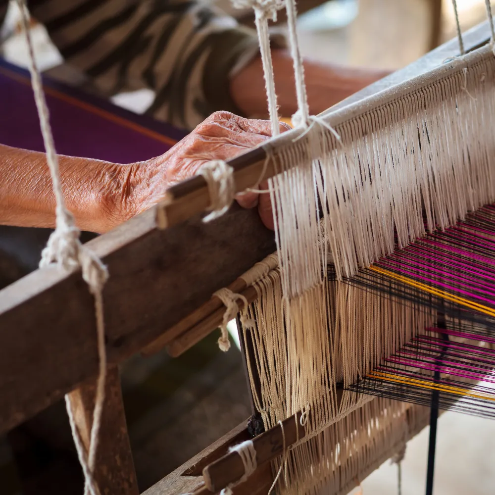 A woman from a Thai hill tribe is hand weaving on a wooden loom.