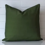 White wall with an olive green outdoor cushion laying against it. It has a distinctive waterproof fabric and has a square shape.