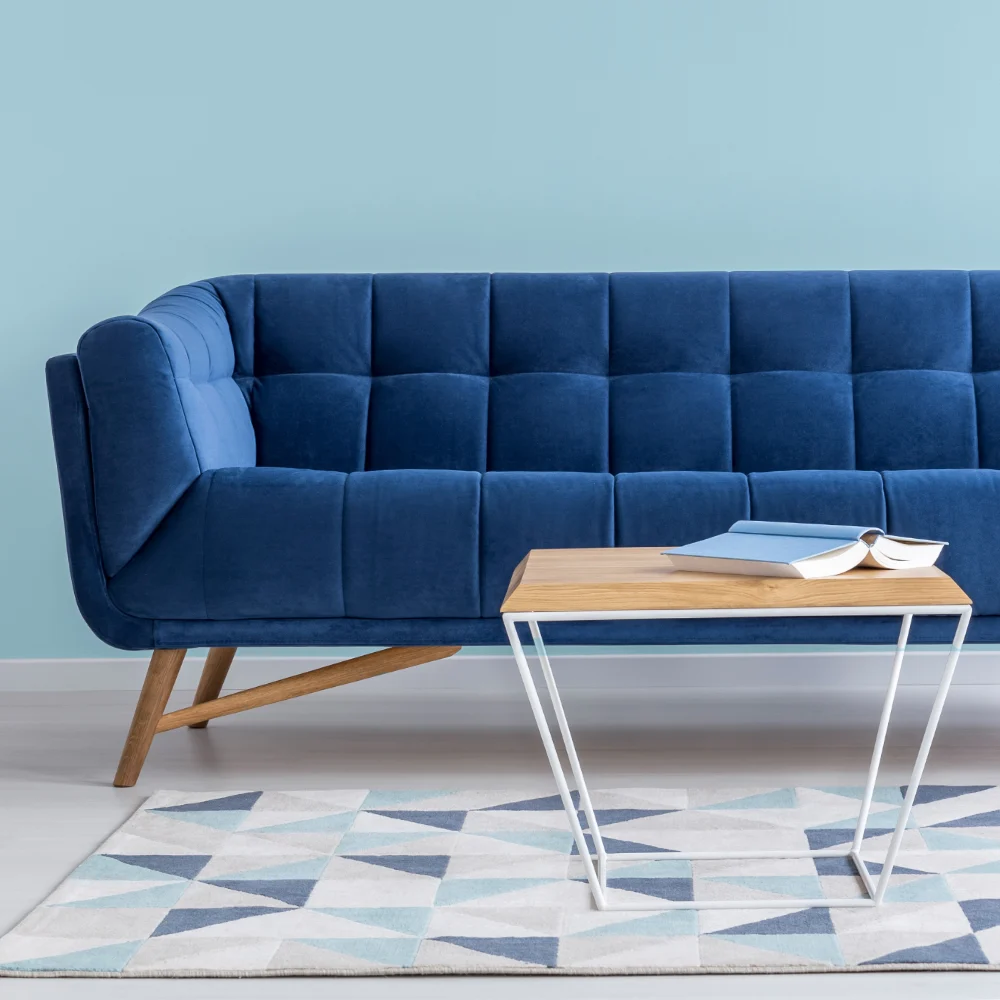 A blue sofa is shown against a light blue wall and behind a small wooden table that rests on a matching multi-coloured rug.